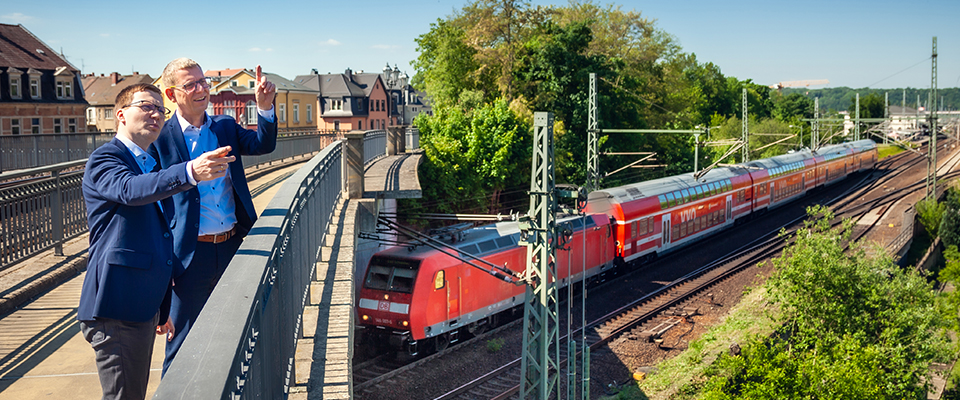 Zwei M&auml;nner stehen auf einer Br&uuml;cke und zeigen auf eine rote S-Bahn, die auf Gleisen neben der Br&uuml;cke f&auml;hrt.